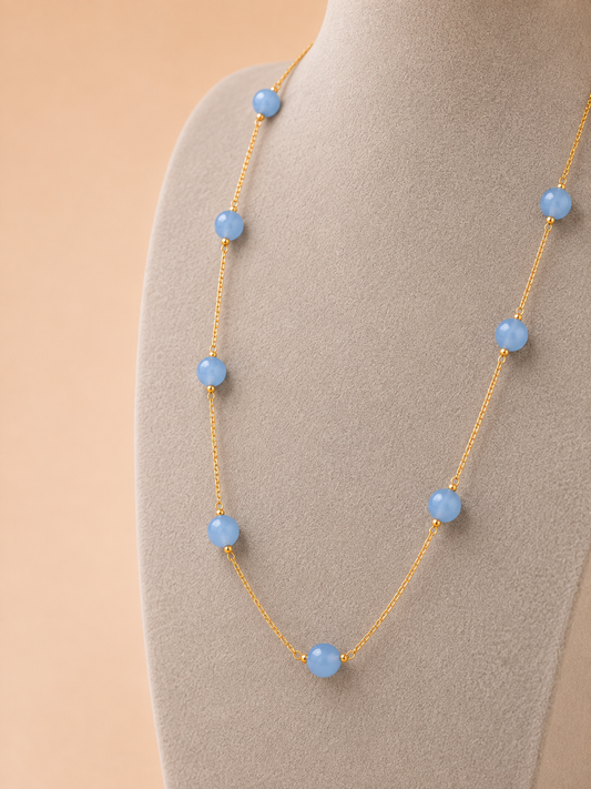 A delicate gold chain necklace with evenly spaced light blue beads, displayed on a neutral beige jewelry bust against a soft peach background.