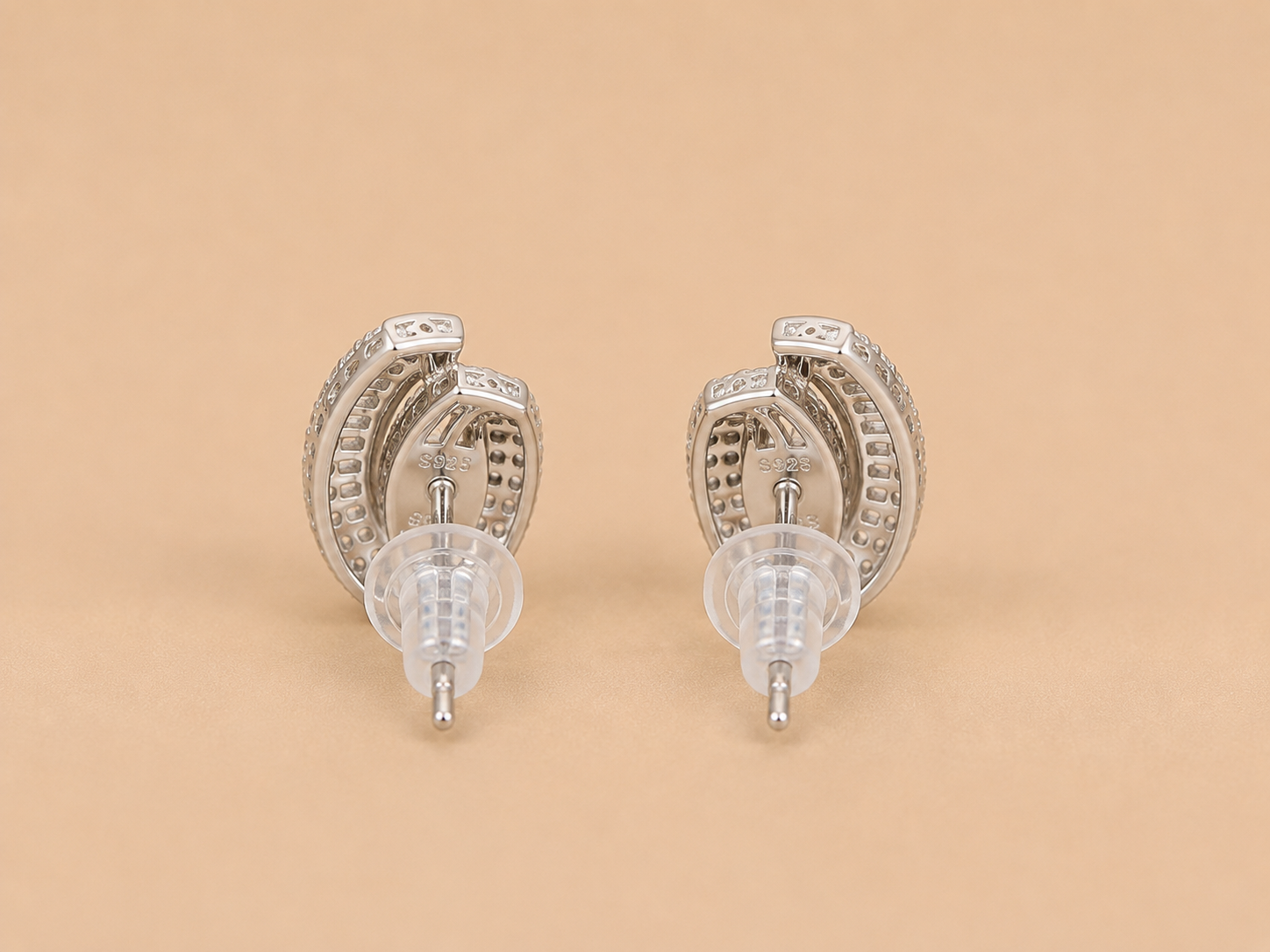 A pair of silver-toned crescent-shaped stud earrings with textured inner bands and clear plastic display tips, shown on a solid beige background.
