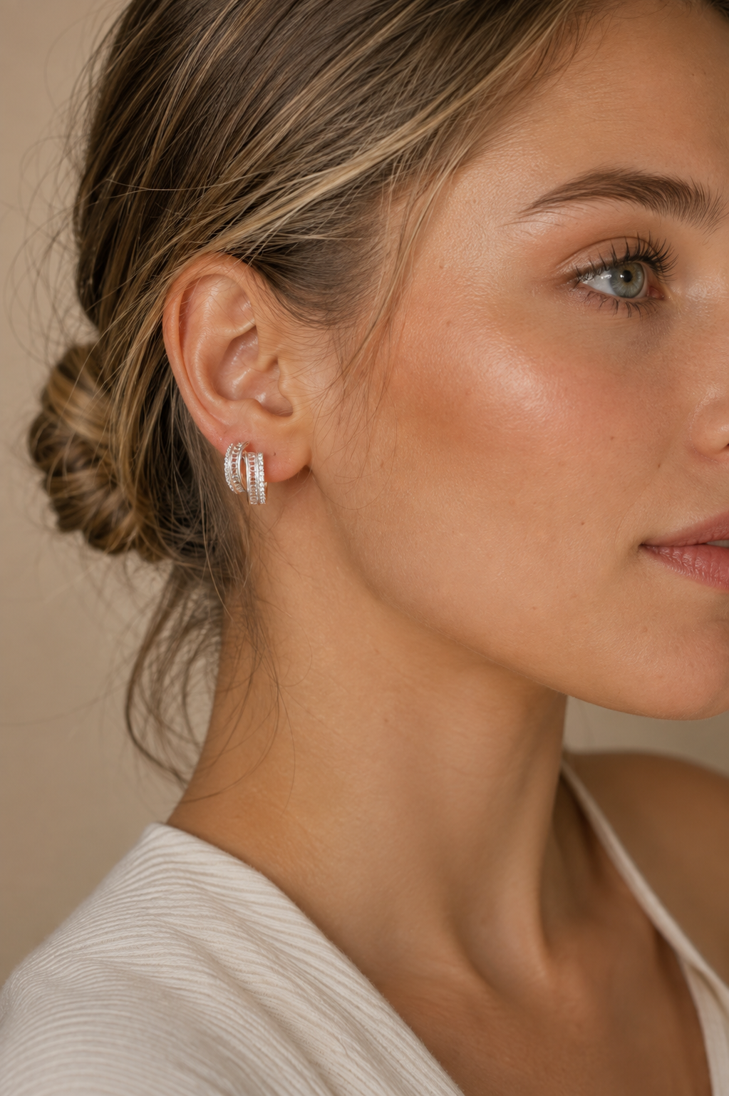 A close-up of a woman wearing a pair of silver hoop earrings adorned with small clear stones, set against a soft beige background.