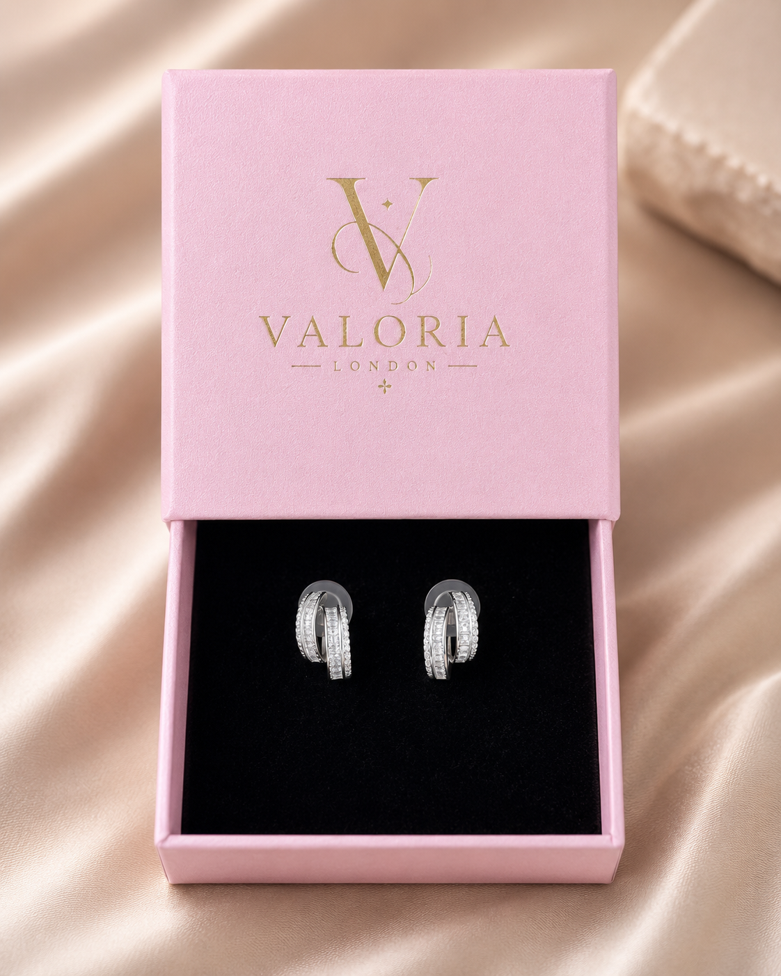 A pair of silver hoop earrings displayed in an open pink Valoria London gift box with a black velvet insert, set against a soft champagne-colored satin fabric background.