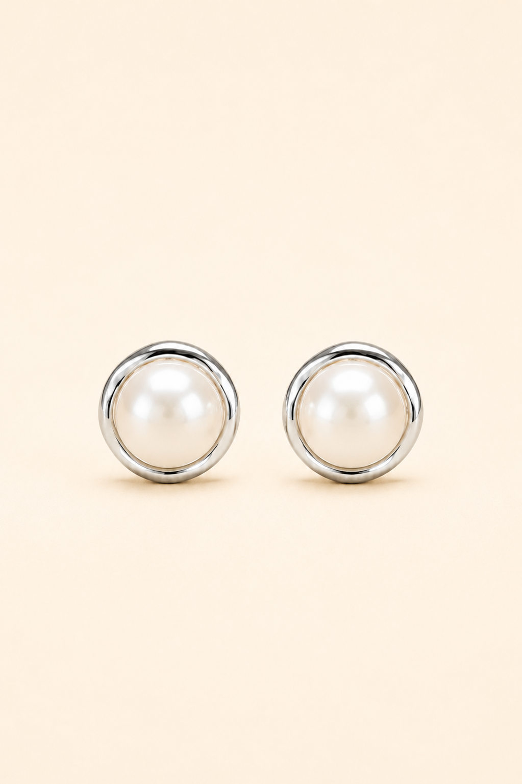 A pair of round stud earrings featuring white faux pearls set in polished silver-tone metal, displayed on a solid light beige background.