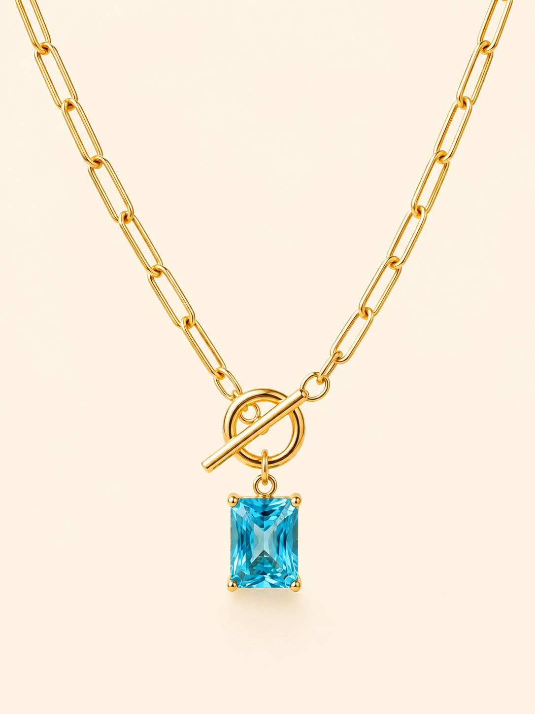 Gold paperclip chain necklace with a toggle clasp and a rectangular light blue gemstone pendant, shown on a light cream background.