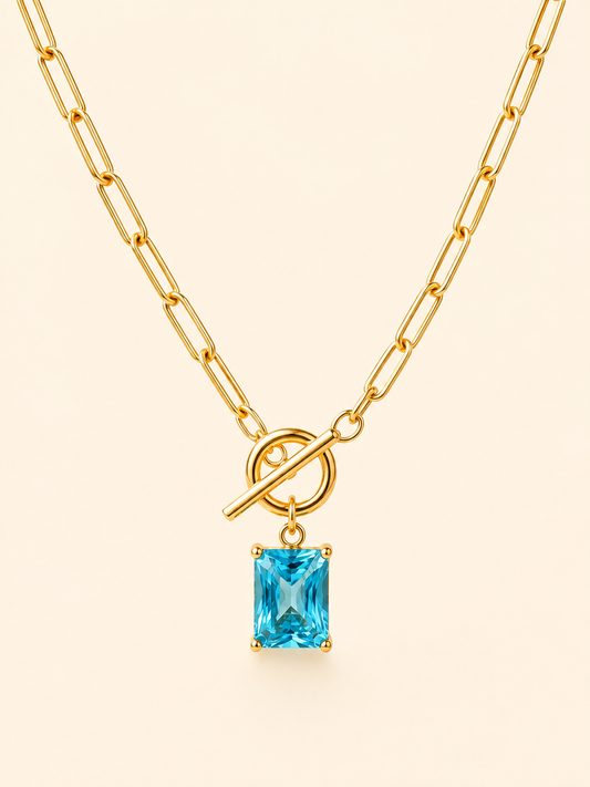 Gold paperclip chain necklace with a toggle clasp and a rectangular light blue gemstone pendant, shown on a light cream background.