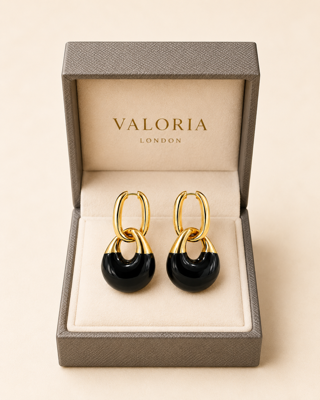 A pair of Valoria London gold hoop earrings with black circular drops, presented in an open textured gray jewelry box with a cream interior.