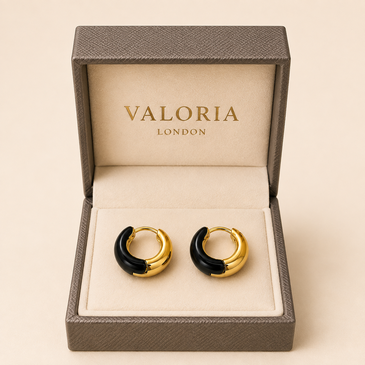 A pair of gold and black crescent-shaped hoop earrings displayed inside an open textured gray Valoria London gift box on a warm beige background.