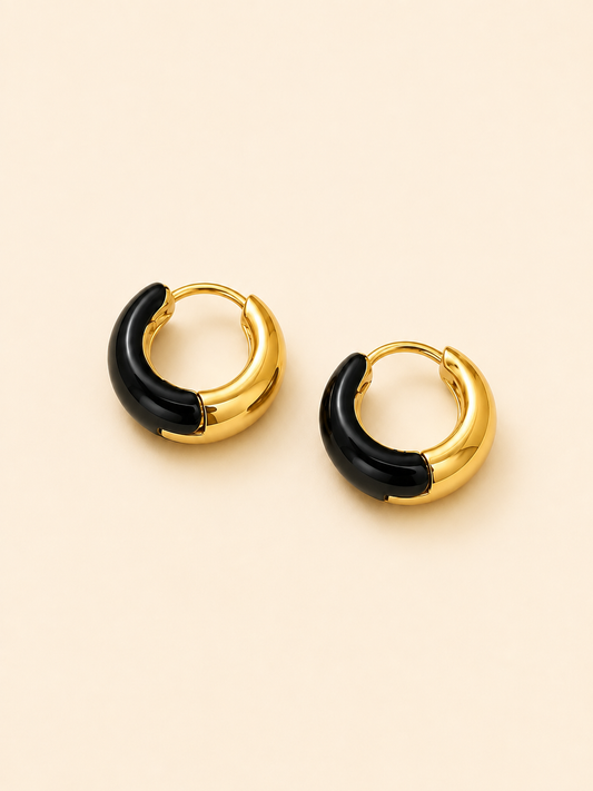 A pair of gold huggie hoop earrings with a glossy black curved inlay, displayed on a warm beige background.