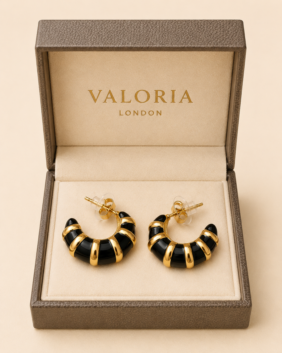 A pair of black and gold crescent-shaped hoop earrings displayed inside an open Valoria London jewelry box on a warm beige background.