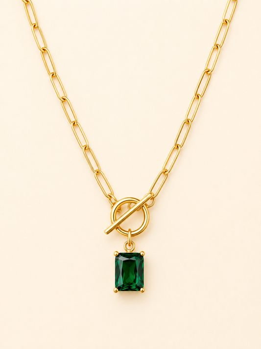 A gold paperclip chain necklace with a toggle clasp and a rectangular emerald-cut green gemstone pendant, shown on a light cream background.
