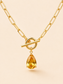 Gold paperclip chain necklace with a toggle clasp and a dangling pear-cut yellow gemstone pendant, shown on a warm beige background.
