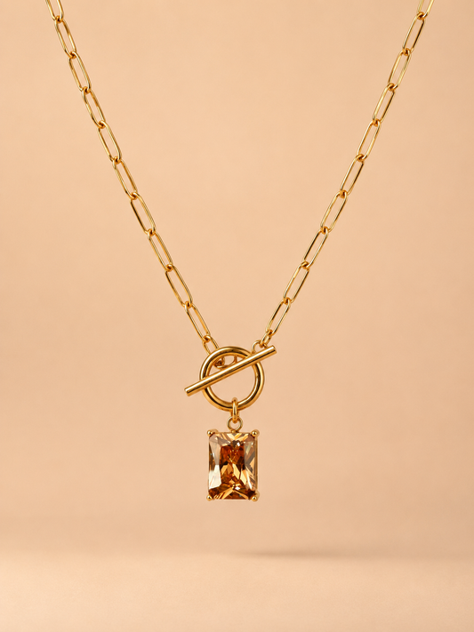 Close-up of gold toggle necklace with rectangular amber crystal pendant on a nude background