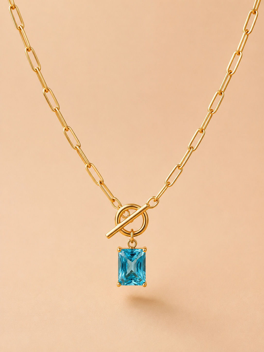 Close-up of a gold toggle necklace with a rectangular blue crystal pendant on a nude background