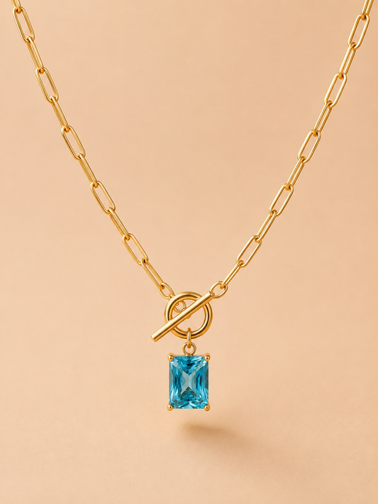Close-up of a gold toggle necklace with a rectangular blue crystal pendant on a nude background