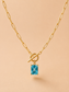 Close-up of a gold toggle necklace with a rectangular blue crystal pendant on a nude background