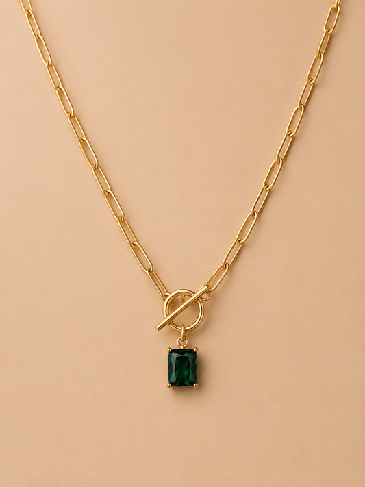 Close-up of gold toggle necklace with rectangular emerald green pendant on a nude background