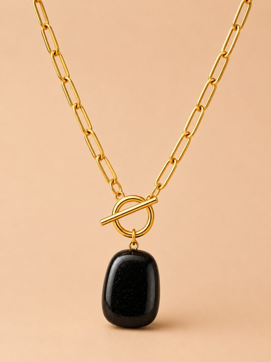 Gold paperclip chain necklace with black stone pendant and toggle clasp on a nude background