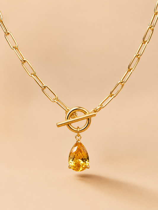 Close-up of a gold toggle necklace with a yellow teardrop crystal pendant on a nude background