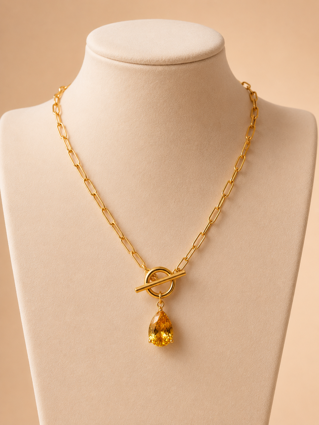 Gold paperclip chain necklace with a yellow teardrop pendant and toggle clasp on a nude background