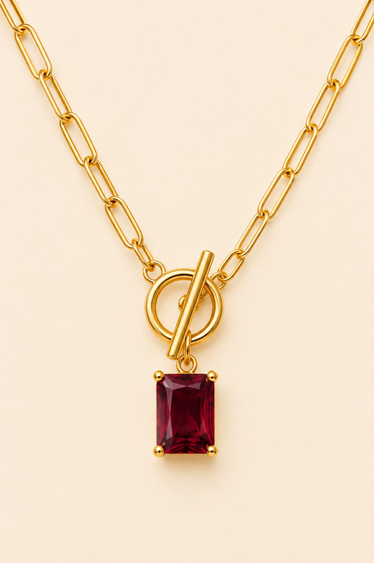 Gold paperclip chain necklace with a deep red rectangular gemstone pendant and toggle clasp on a soft ivory cream background.