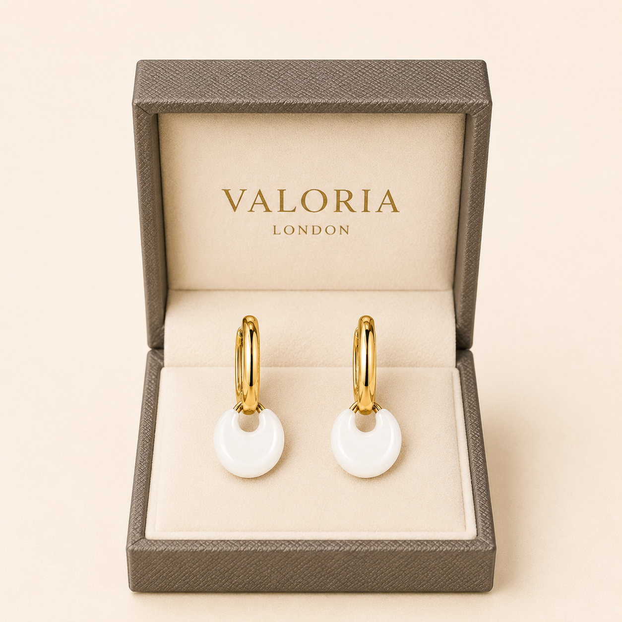 A pair of gold hoop earrings with round white disc pendants, presented in an open Valoria London jewelry box on a warm beige background.