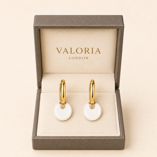 A pair of gold hoop earrings with round white disc pendants, presented in an open Valoria London jewelry box on a warm beige background.