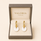 A pair of gold hoop earrings with round white disc pendants, presented in an open Valoria London jewelry box on a warm beige background.
