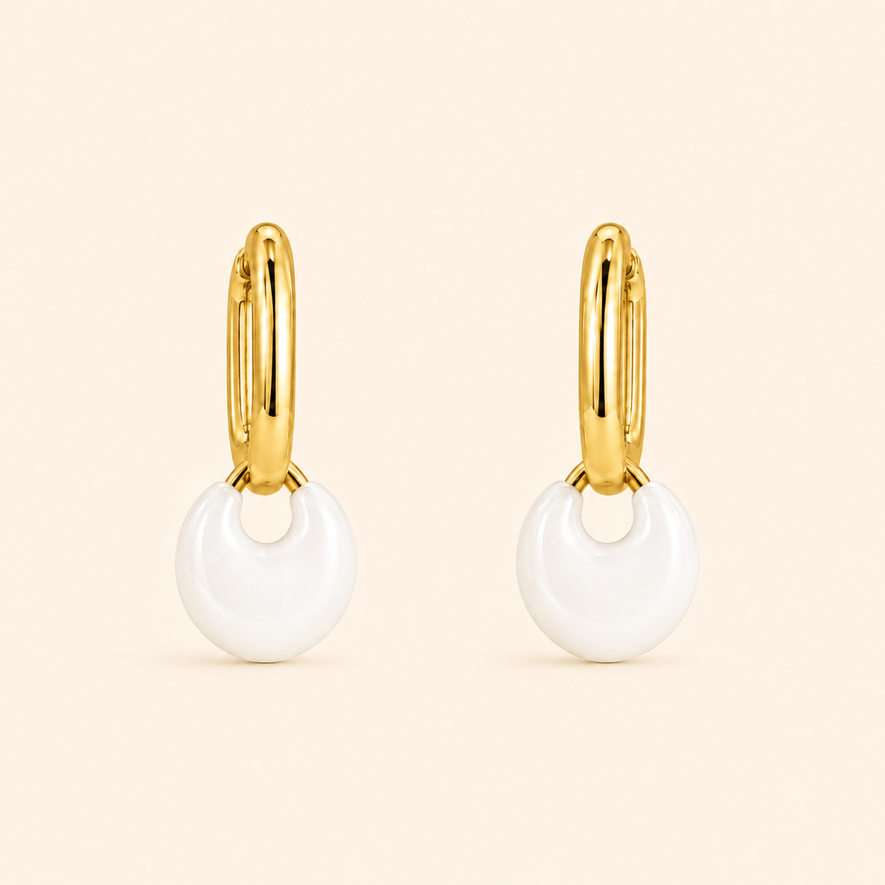 A pair of gold huggie hoop earrings with a small white crescent-shaped drop charm, shown on a solid pale beige background.