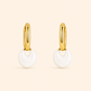 A pair of gold huggie hoop earrings with a small white crescent-shaped drop charm, shown on a solid pale beige background.