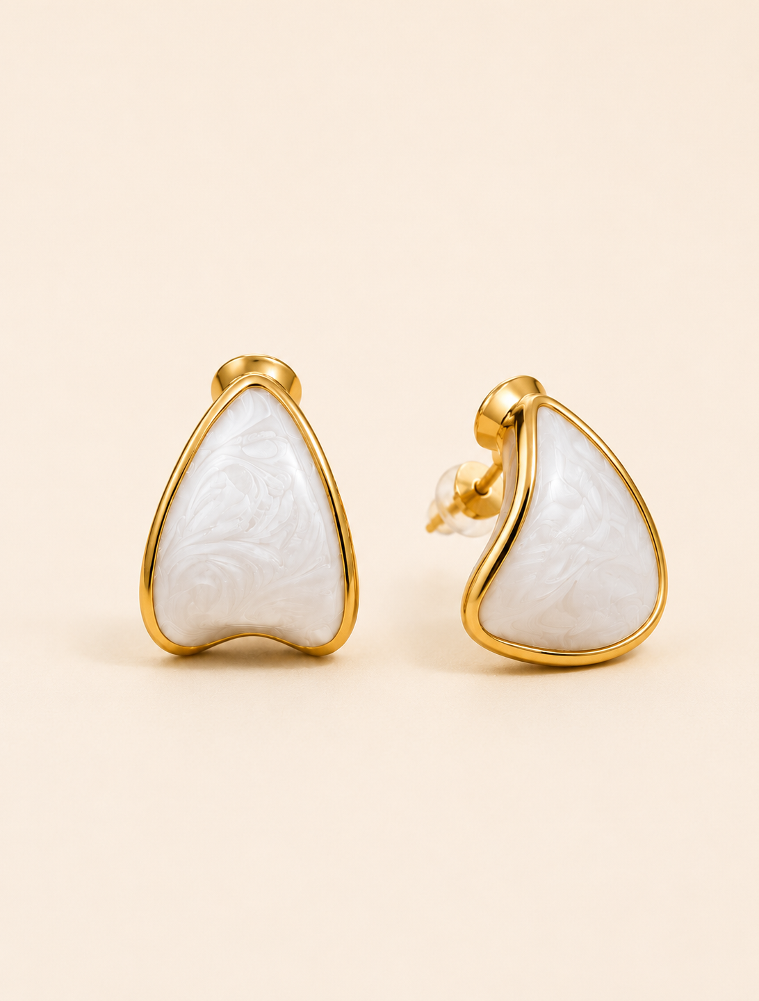 A pair of gold stud earrings with teardrop-shaped white carved stone inlays, displayed on a warm beige background.