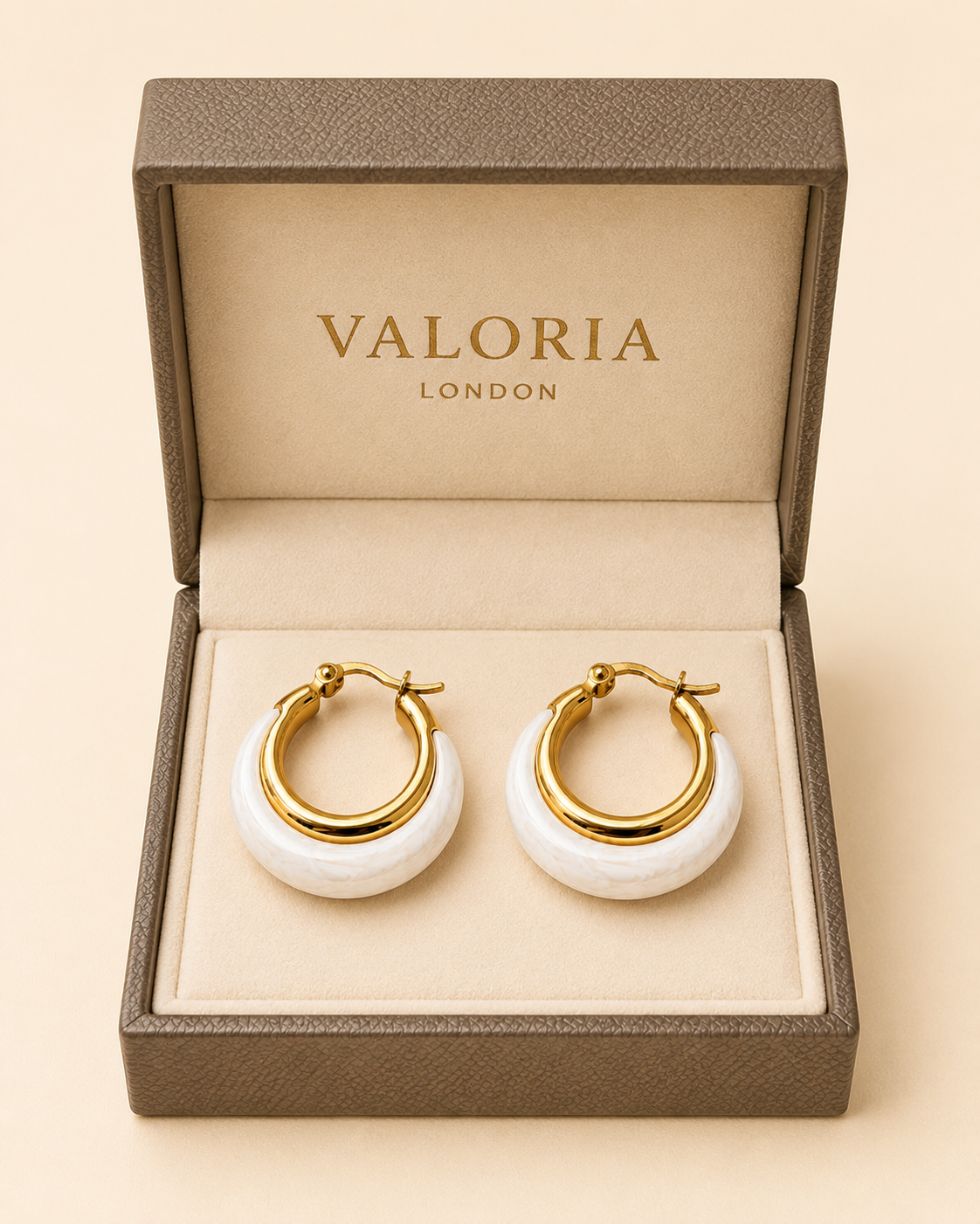 A pair of Valoria London gold hoop earrings with white stone inlays, presented in an open textured brown jewelry box on a beige background.