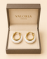 A pair of Valoria London gold hoop earrings with white stone inlays, presented in an open textured brown jewelry box on a beige background.