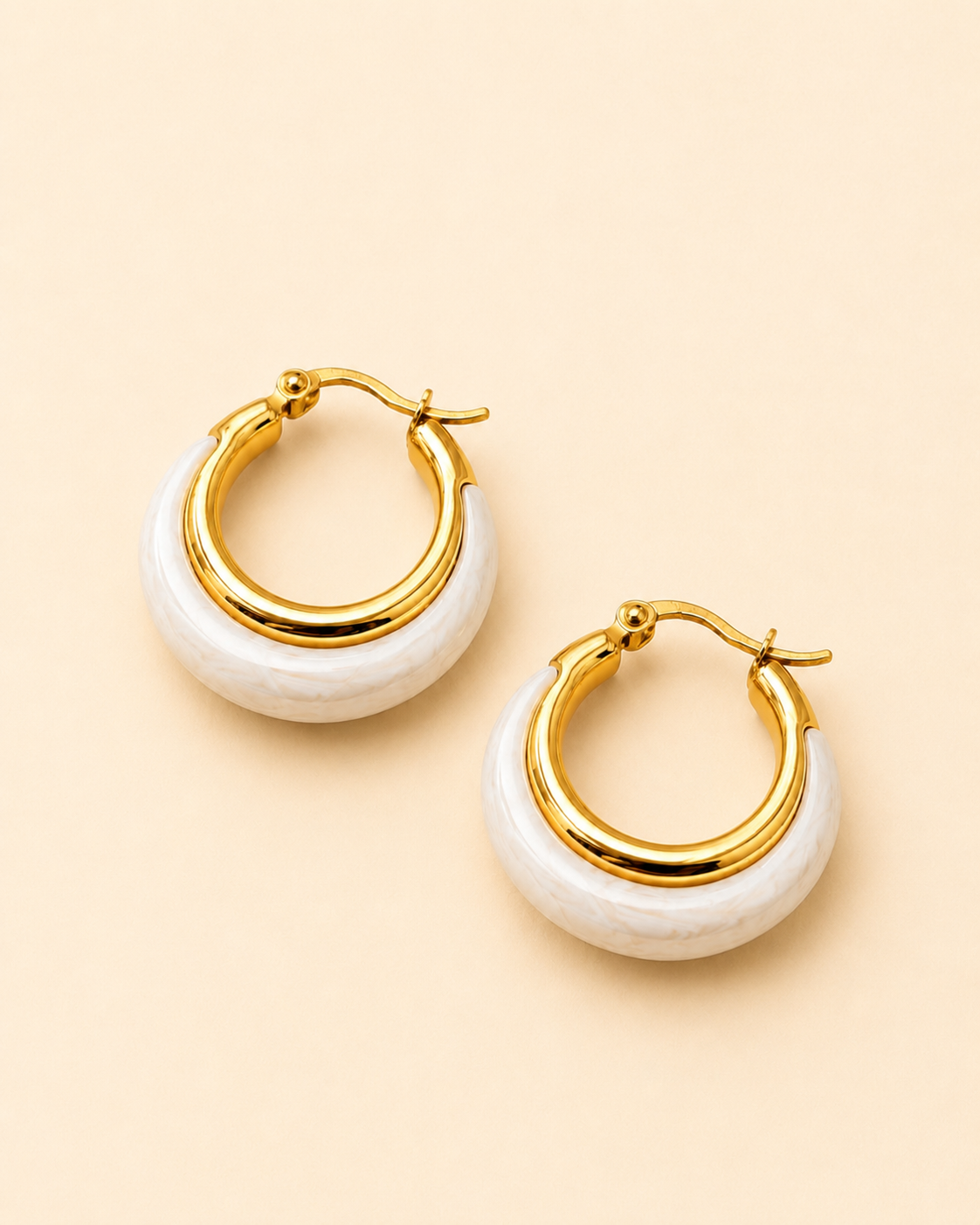 A pair of gold hoop earrings with a white textured crescent-shaped inlay, displayed on a solid pale peach background.