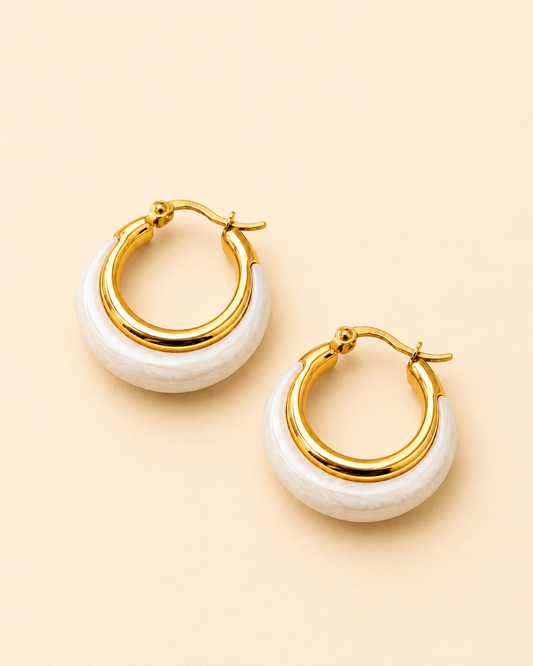A pair of gold hoop earrings with a white textured crescent-shaped inlay, displayed on a solid pale peach background.